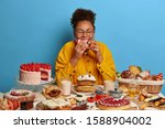 Small photo of Cheat meal and gluttony concept. Ethnic curly woman eats strawberry creamy cake with much calories, has sweet lunch, tastes various desserts, leads sweet life with confectionery. Female fan of bakery
