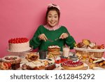 Cheerful mixed race woman eats with big appetite creamy pancakes, has unhealthy diet, looks gladfully, being sweet tooth, overeats desserts, isolated over pink background, cannot prevent obesity