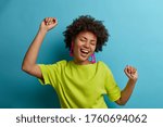 I am winner! Ecstatic overjoyed African American woman dances carefree, celebrates victory and success, dressed in green casual t shirt, feels lively and energetic, isolated on blue background