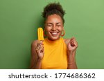 Happy dark skinned millennial girl eats yellow ice cream of mango flavor, clenches fist with anticipation of something awesome happen, has toothy smile, wears bright summer clothes, isolated on green