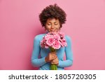 Attractive pleased dark skinned model receives flowers as present, stands with closed eyes, enjoys her favorite gerberas, isolated on pink background. Romantic portrait. Woman gets gift from husband