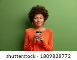 Studio shot of satisfied dark skinned girl with Afro hair holds paper cup of hot coffee, closes eyes, wears orange jumper, green background. Beautiful young woman enjoys aromatic cappuccino.