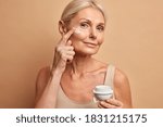Close up shot of middle aged beautiful woman applies anti aging cream on face undergoes beauty treatments cares about skin poses against beige background. Wrinkled female model with cosmetic product