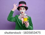 Photo of shocked mad hatter being on tea party wears big hat lace gloves and green costume poses with beverage has professional bright makeup isolated on purple background. Halloween carnival