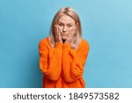 Small photo of Bored displeased woman looks sadly at camera keeps hands on cheeks being sick and tired of constant reproaches dressed in knitted sweater models against blue background has moody expression.