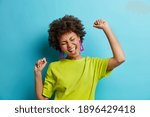 Small photo of Overjoyed African American woman makes winning gesture expresses happiness raises arms and dances carefree celebrates victory dressed in t shirt isolated over blue background. Female triumphant
