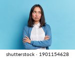 Small photo of Portrait of displeased offended young Asian woman with dark hair keeps arms folded looks angrily aside doesnt agree with somebodys opinion wears neat clothes isolated over blue studio background