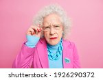Small photo of Portrait of scrupulous granny has attentive gaze at camera bad eyesight keeps hand on rim of spectacles dressed in fashionable clothes always cares about her appearance poses indoor. Old style concept