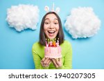 Portrait of cheerful beautiful Asian woman blows candles on birthday cake wears unicorn headband makes wish has happy festive mood isolated over blue background clouds white above. Festivity concept