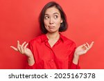 Small photo of Clueless hesitant beautiful Asian woman with short dark hair spreads palms sideways shrugs shoulders has indecisive expression dressed in shirt stands against vivid red background. Who knows.
