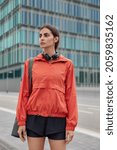 Small photo of Vertical shot of thoughtful sporty woman dressed in anorak and shorts carries yoga mat goes to gym poses on road against city buildings concentrated away listens music while having training.