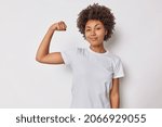 Small photo of Confident woman feels strength and power raises arm flexes biceps and looks proud of her own achievements has strong muscles wears casual t shirt isolated over white background brags with fit body