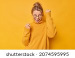 Small photo of Yes finally success. Positive woman raises arms with clenched fists celebrates something has upbeat mood feels euphoric wears round spectacles and casual jumper poses against bright yellow background