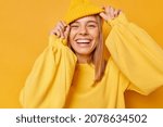 Small photo of Happy playful young woman smiles broadly shows white teeth keeps hands on hat dressed in casual jumper expresses sincere emotions being in good mood isolated over yellow background. Joy concept