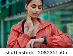 Cropped shot of serious woman checks pulse on neck monitors fitness activity has quick heart rate wears red jacket poses outdoors against blurred background controls her health. Devices for sport