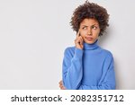 Small photo of Photo of serious young curly woman keeps finger on temple tries to concentrate considers something contemplates about important thing wears casual blue jumper isolated over white background.