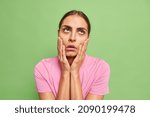 Small photo of Uninterested bored young woman keeps hands on cheeks fed up of dull conversation looks above makes facepalm and rolls eyes hears nonsense wears casual t shirt isolated over green background.