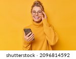 Small photo of Positive woman smiles happily keeps hand on rim of spectacles feels cheerful wears comfortable sweater downloads cool application edits pics poses against yellow background. Technology concept