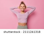 Horizontal shot of female athlete takes break after cardio workout keeps hands behind head smiles broadly stays in good physical shape wears cropped top and leggings isolated over pink background