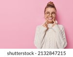 Small photo of Horizontal shot of pretty cheerful woman with bun hairstylepoints index fingers at smile shows white teeth concentrated away wears spectacles and white knitted sweater isolated on pink background