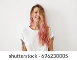 Horizontal portrait of pleasant-looking Caucasian female with long hair, pink on tips, having tattooes on arms, wearing white casual T-shirt, covering her face with hair, looking happily in camera