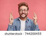 Glad positive bearded man crosses fingers, closes eyes with pleasure, anticipate hearing good news, isolated pink background. Happy male rejoices that his dreams came true. Excitement concept