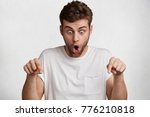 Photo of shocked bearded male looks down with great disbelief, expresses surprisment and astonishment, indicate with fore fingers at floor, isolated over white concrete wall. Caucasian man gestures