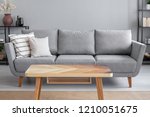 Wooden table and big grey couch with pillows in living room of trendy apartment, real photo with copy space on the wall