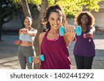 Portrait of young curvy woman with friends exercising with dumbbells. Group of three multiethnic girls working out in park during sunset. Smiling women in sportswear using dumbbell to lose weight.