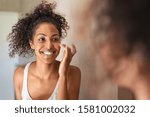 Young black woman applying moisturising cream on cheeck while standing in front of the mirror in bathroom. African american girl applying face cream. Beauty hydrating moisturizer and skincare routine.