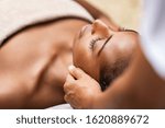 Beautiful african woman getting face massage in beauty spa. Black girl with closed eyes relaxing in outdoor spa while getting head massage. Serene woman relaxing outdoor in a beauty center.