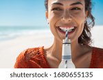 Closeup face of young woman drinking fresh sparkling water from a glass bottle at beach. Portrait of beautiful carefree girl drinking soft drink with straw during summer. Smiling girl staying hydrated