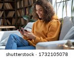 Happy young hispanic latin teen girl sit on sofa at home holding phone looking at screen watching social media video content, movie or stream, video calling online in mobile app using smart phone.