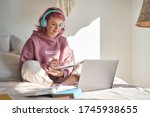 Hipster teen girl school student with pink hair wear headphone write notes watch video online webinar learn on laptop sit in bed distance elearning course video conference pc call in bedroom at home.
