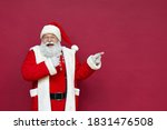 Funny happy excited old bearded Santa Claus face wearing costume looking at camera showing pointing fingers aside advertising Christmas promotion, New Year xmas discount ad isolated on red background.