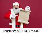 Happy excited old bearded Santa Claus wearing costume holding Merry Christmas wishlist paper roll pointing finger at blank empty xmas wish list letter standing isolated on red background, copy space.