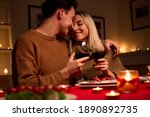 Happy young couple in love hugging holding glasses, drinking wine, celebrating Valentines day dining at home together, having romantic dinner date with candles sitting at table, embracing and bonding.
