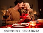 Happy young couple celebrating anniversary or Valentines day having romantic dinner at home table. Loving man giving red gift box hugging beloved woman making present surprise on date in candle light.