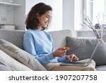 Happy hispanic young woman consumer holding credit card and laptop buying online at home. Female shopper customer shopping on ecommerce website market place making digital payment using bonus money.