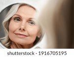 Happy 50s middle aged woman model touching face skin looking in mirror reflection. Smiling mature old lady pampering, healthy moisturized skin care, aging beauty, skincare treatment cosmetics concept.
