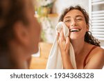 Young woman wiping her face with towel after waking up in the morning. Beautiful happy smiling girl holding towel near facial skin after washing face. Happy woman cleaning and drying skin with napkin.