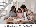 Small photo of Excited indian family with child daughter unpack parcel at home. Happy parents and teen kid daughter open postal box receive present gift in online shopping delivery package sitting on sofa together.