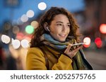 Happy woman recording message using voice on smartphone. Happy woman recording audio on mobile phone in the street at evening. Smiling woman speaking on her smart phone in winter evening.