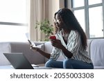 Small photo of Serious young black African woman holding paper document calculating rent or money savings, paying bills in mobile application on cell phone doing monthly paperwork sitting on couch at home.