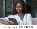 Young black African woman wearing glasses holding digital tablet sitting on couch at home learning online, watching videos, surfing internet, doing e shopping using technology device relaxing on sofa.