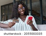 Small photo of Happy young black African woman customer shopper holding credit card using cell phone mobile app buying fashion clothes paying online making purchase in ecommerce digital store on smartphone at home.