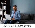 Successful mature indian businessman sitting on couch typing on laptop with wireless earphones. Mixed race businessman sitting on couch while working from home during video call. Business man working 