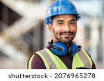 Portrait of satisfied construction site manager wearing safety vest and blue helmet with copy space. Young middle eastern architect watching construction site with confidence and looking at camera.