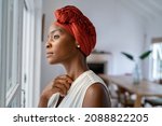 Small photo of Pensive woman wearing headscarf looking outside window in contemplation. Mature black woman wearing a traditional turban and thinking near window at home. Worried african mature woman with cancer.
