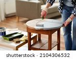 furniture renovation, diy and home improvement concept - close up of woman in gloves with paint roller painting old wooden table in grey color